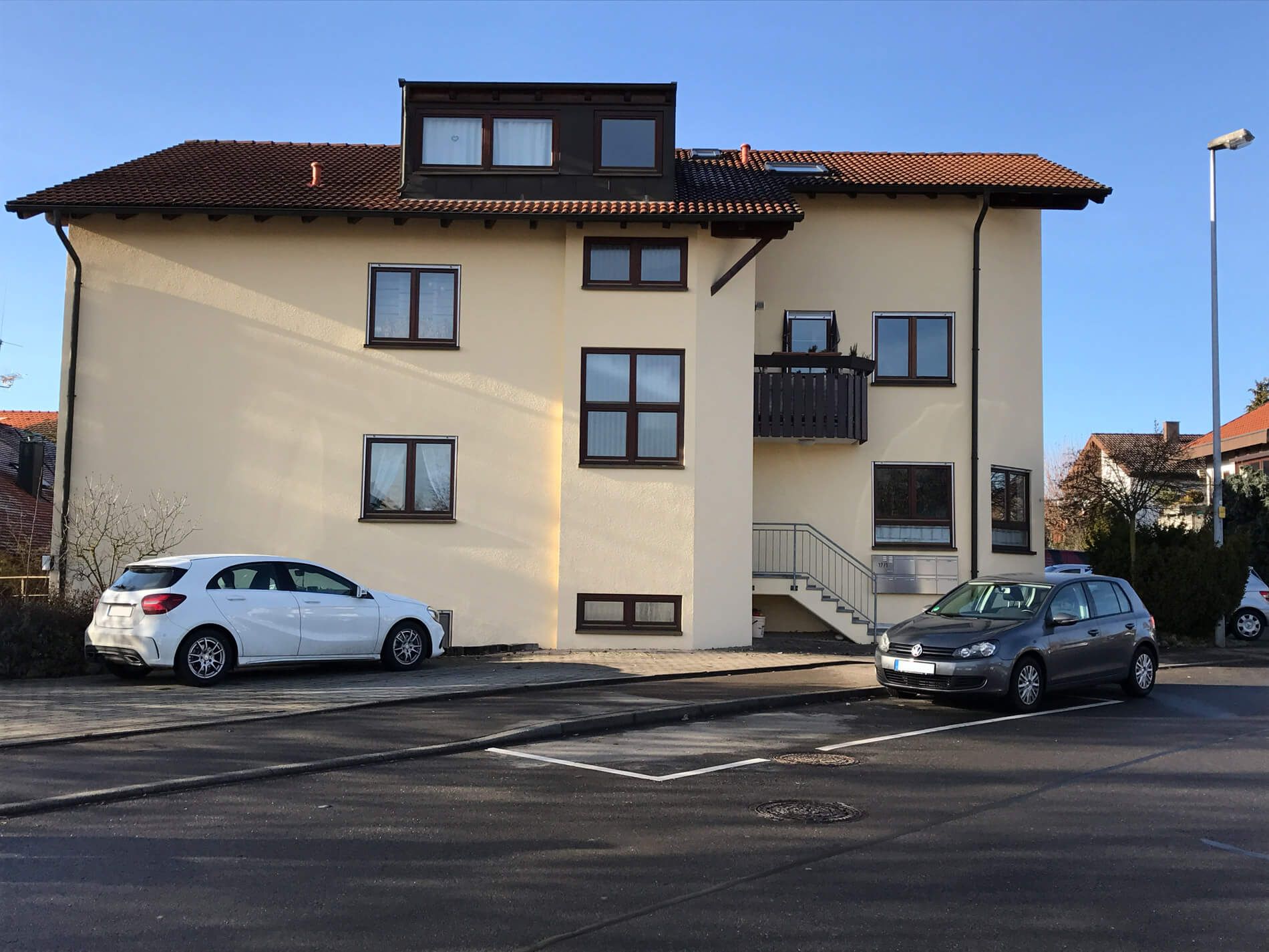 Mehrfamilienhaus mit rotbraunem Satteldach, gelber Putzfassade und dunklen Fensterrahmen, Parkplatz im Vordergrund