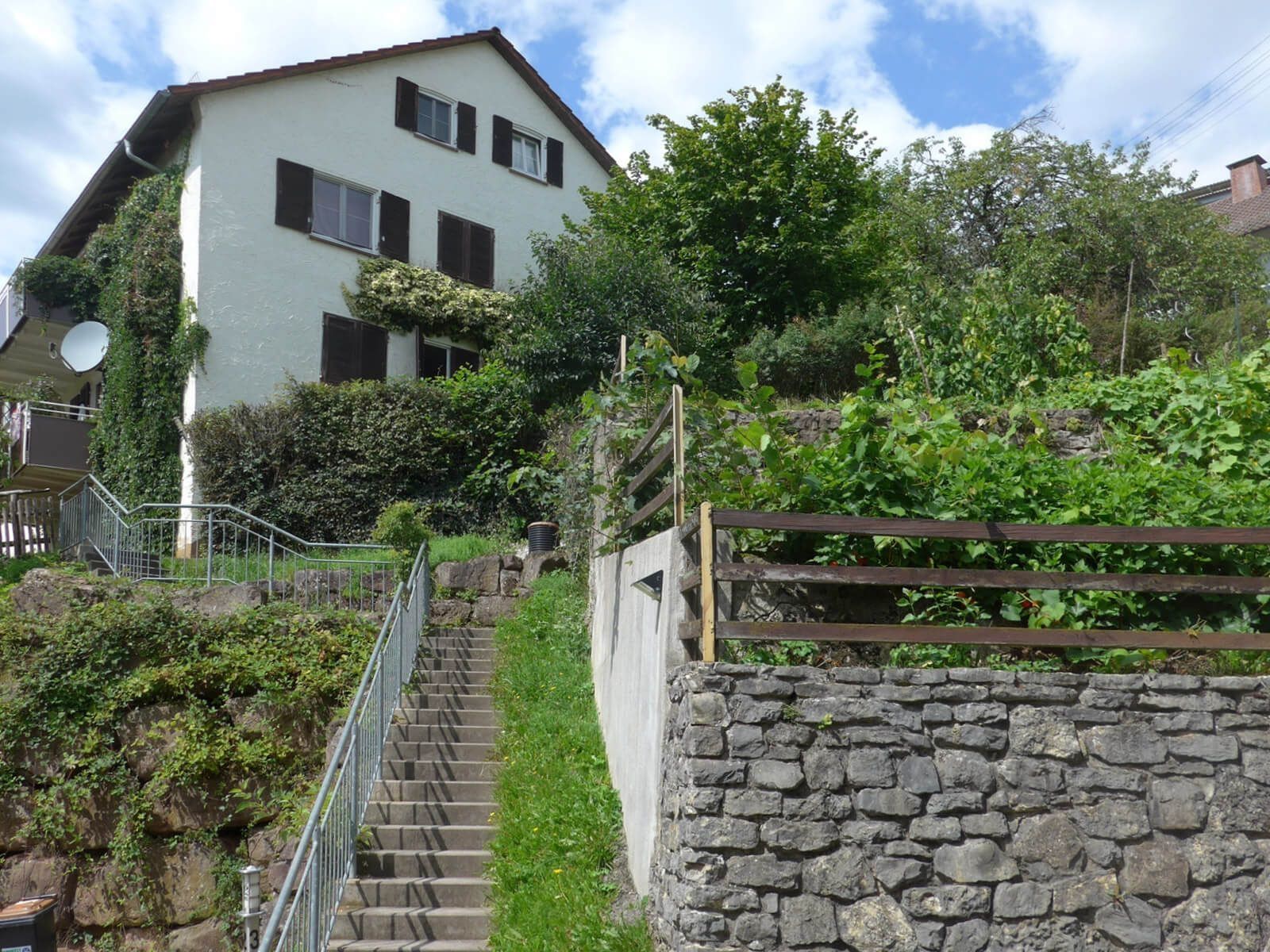 Mehrfamilienhaus in Hanglage mit heller Putzfassade, Treppenaufgang und üppig begrüntem Garten